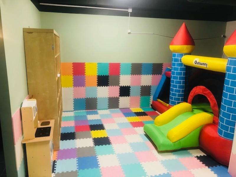 Lekrum för barn med färgglada skummattor på golvet, ett lekkök i trä till vänster och en uppblåsbar hoppborg med rutschkana till höger.