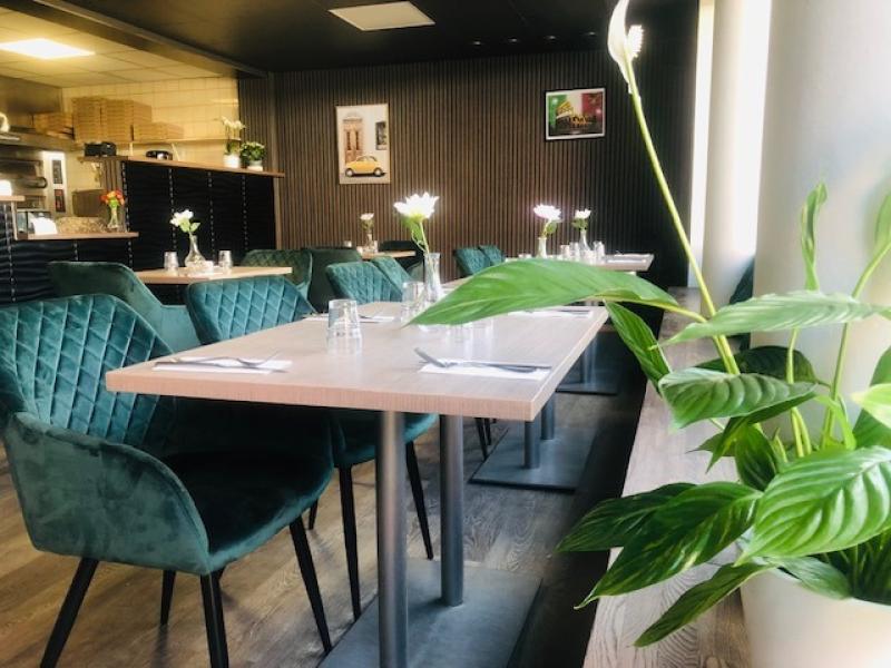 Modern restauranginredning med gröna stoppade stolar, träbord med glas och blommor samt en grön växt vid fönstret.