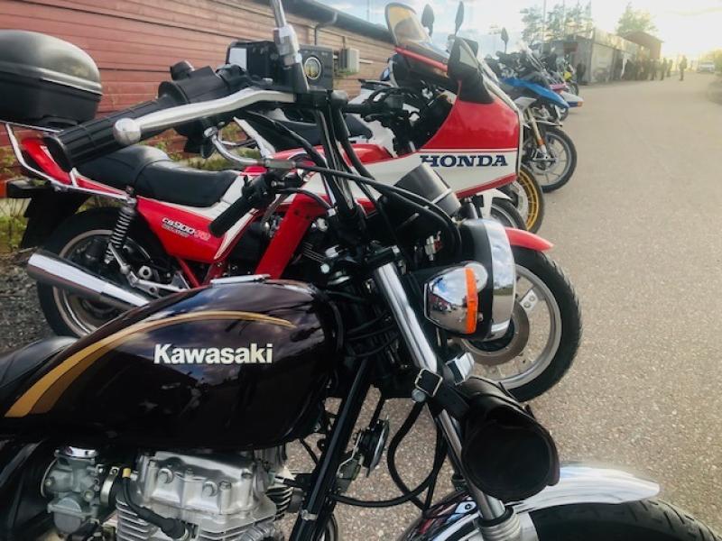 Flera motorcyklar, bland annat en svart Kawasaki och en röd Honda, står parkerade på rad längs en asfalterad gata intill en träbyggnad.