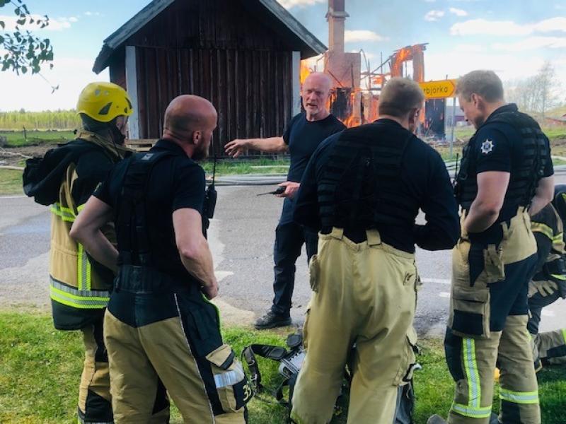 En grupp brandmän i utrustning står och lyssnar på en man medan en träbyggnad brinner i bakgrunden.