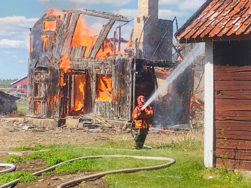 En brandman i skyddsutrustning sprutar vatten på en brinnande träbyggnad, med lågor och rök som stiger upp från byggnaden.