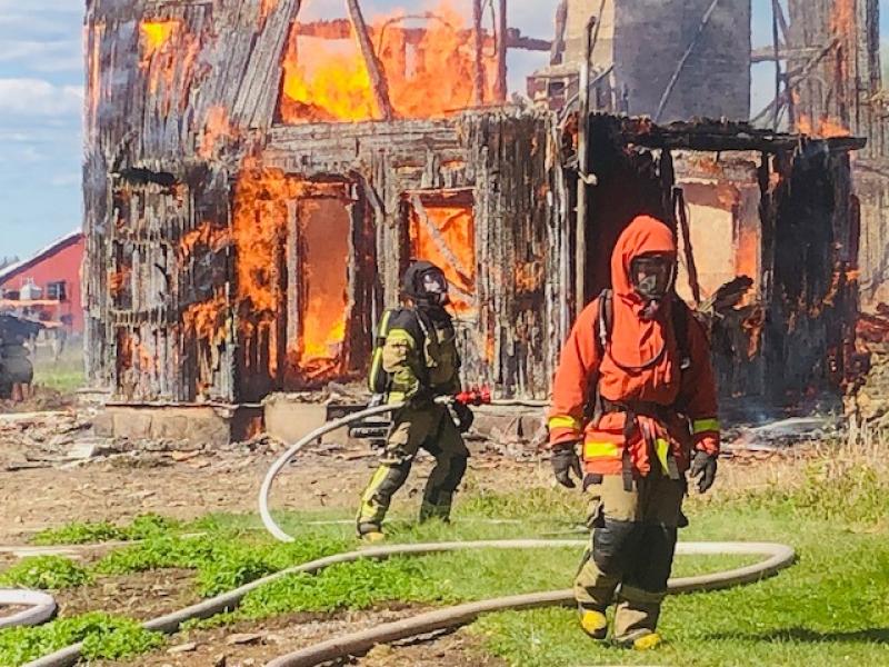 Två brandmän i skyddsutrustning arbetar med att kontrollera en brand som slukar en träbyggnad, med lågor och rök synliga i bakgrunden.