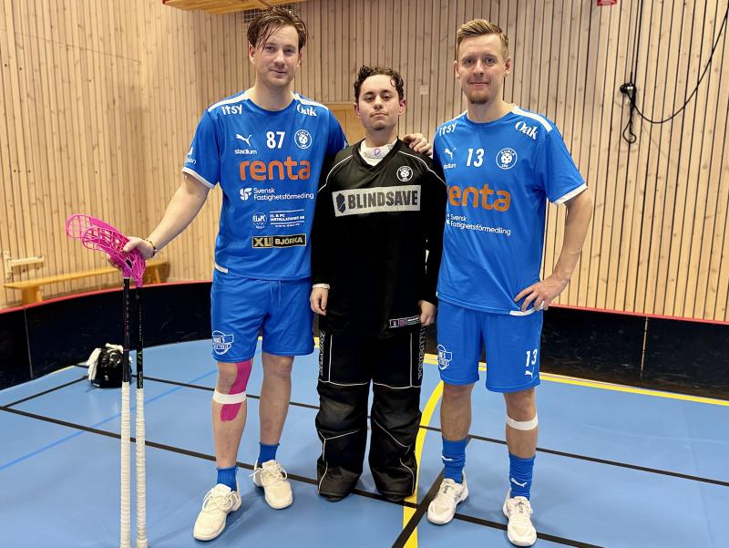 Tre innebandyspelare poserar inomhus; två i blåa uniformer med klubbor, en i svart målvaktsutrustning, mot en träväggsbakgrund.