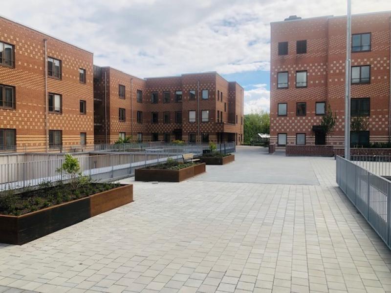 En asfalterad innergård med upphöjda trädgårdsbäddar, omgiven av flervåningshus i tegel under en delvis molnig himmel.