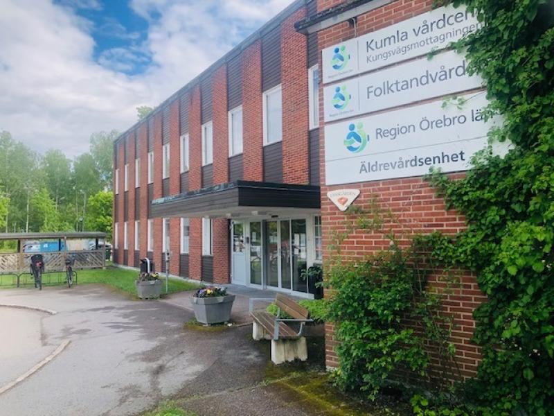 En vårdbyggnad i tegel med skyltar för Kumla vårdcentral, Folktandvården, Region Örebro län och Äldrevårdsenheten vid entrén, omgiven av grönska.
