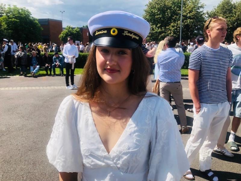 Ung kvinna i vit klänning och studentmössa står utomhus bland en folkmassa en solig dag.