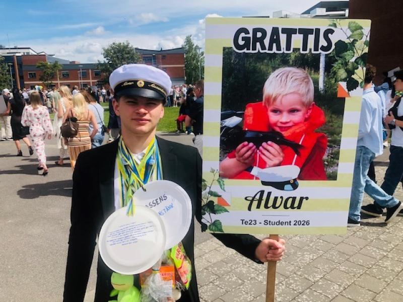 En ung man med studentmössa och medaljer står utomhus och håller i en gratulationsskylt med ett barndomsfoto märkt 