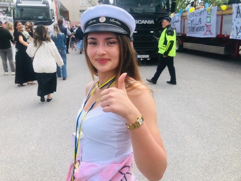En ung kvinna i vit klänning och studentmössa ger tummen upp vid ett utomhusevenemang med människor, lastbilar och banderoller i bakgrunden.