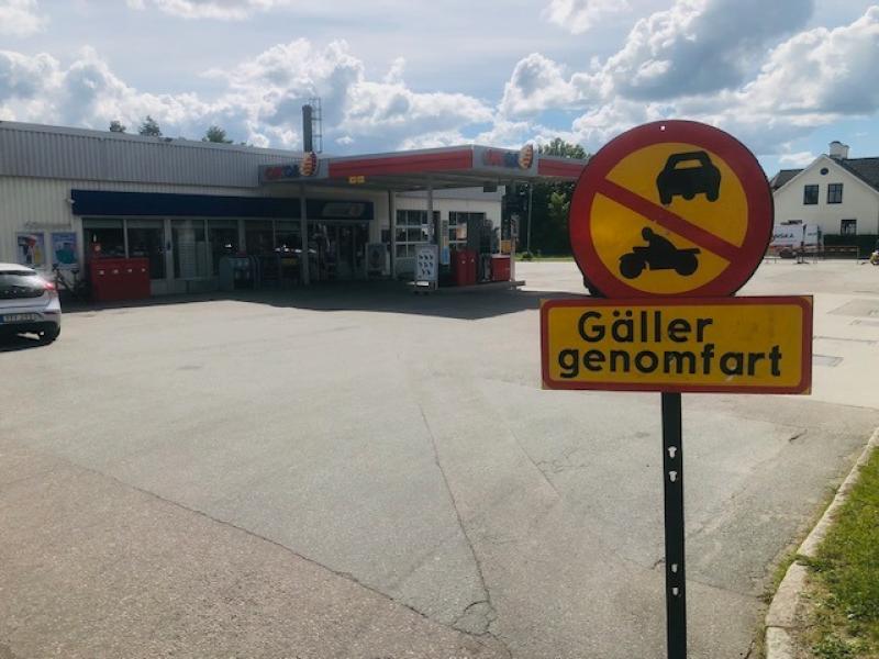 En skylt som förbjuder tillträde för bilar och motorcyklar står framför en bensinstation en solig dag, med en byggnad och parkerade bilar i bakgrunden.