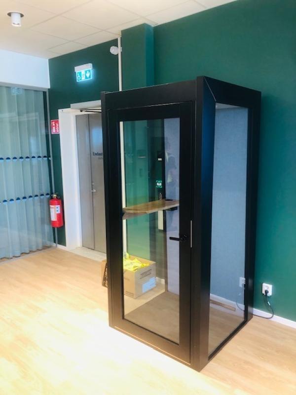 En svart telefonkiosk med glasväggar står i ett kontorsutrymme med ljust trägolv och gröna väggar; inuti kiosken står en kartong.