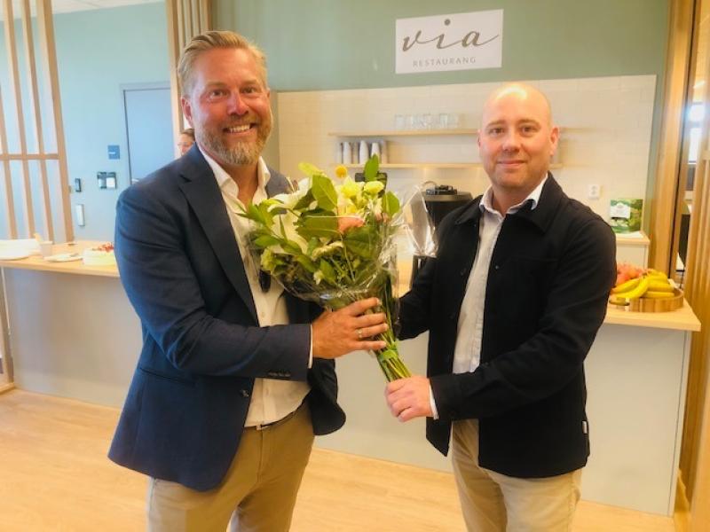 Två män står inomhus; den ene räcker en blomsterbukett till den andre. Båda ler. I bakgrunden syns en disk med frukt och en skylt med texten ”Via Restaurang”.