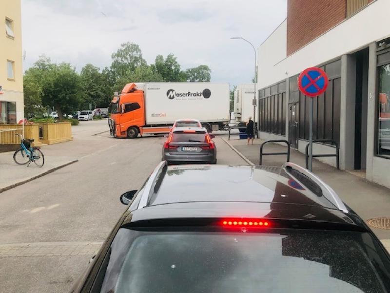 Bilar står och väntar på gatan medan en orange lastbil med vit släpvagn blockerar korsningen; en parkeringsförbudsskylt syns och en person går förbi i närheten.
