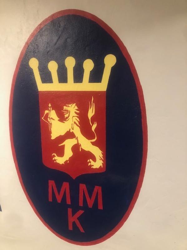 Ett ovalt emblem med ett gul kronat lejon på en röd sköld, med bokstäverna ”MMK” i rött nedanför, allt mot en mörkblå bakgrund.