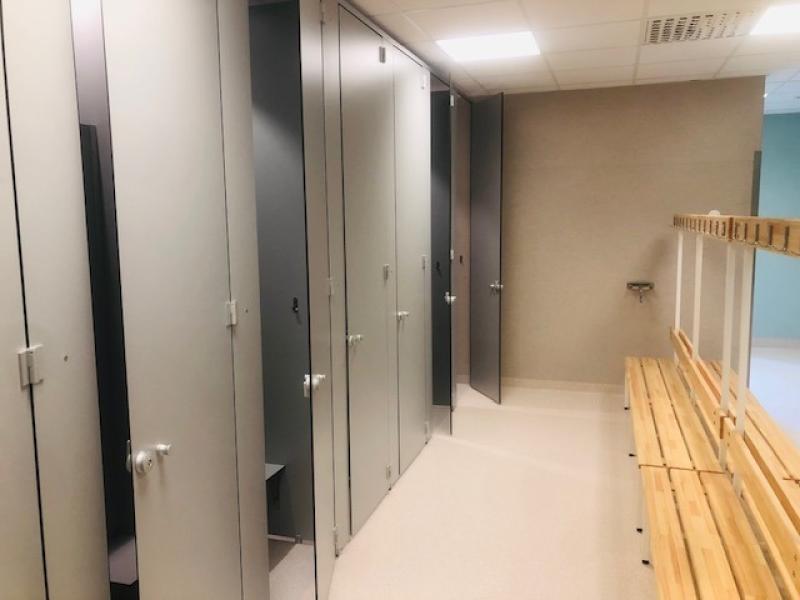 Ett rent omklädningsrum med stängda grå omklädningsbås till vänster och träbänkar med klädkrokar till höger under starka taklampor.