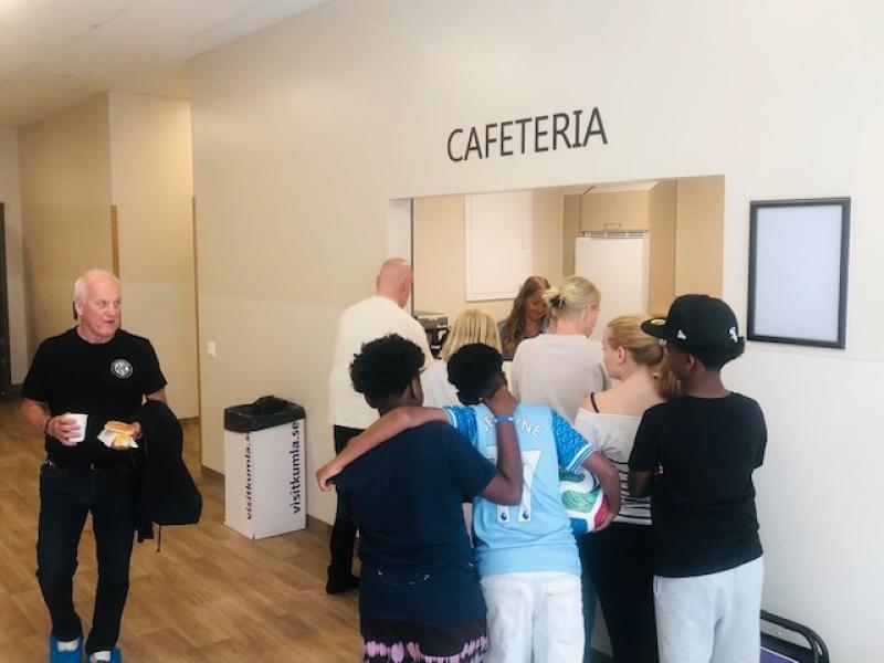 En grupp människor, däribland barn och vuxna, står i kö vid cafeteriadisken. En äldre man går förbi med en kaffe och en smörgås i handen.
