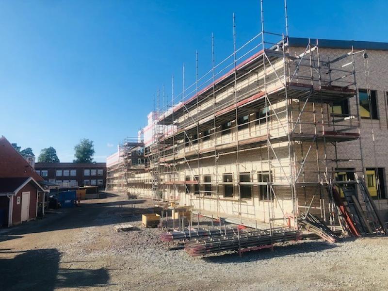 En flervåningsbyggnad under uppförande med byggnadsställningar längs fasaden, omgiven av byggmaterial och utrustning en klar dag.
