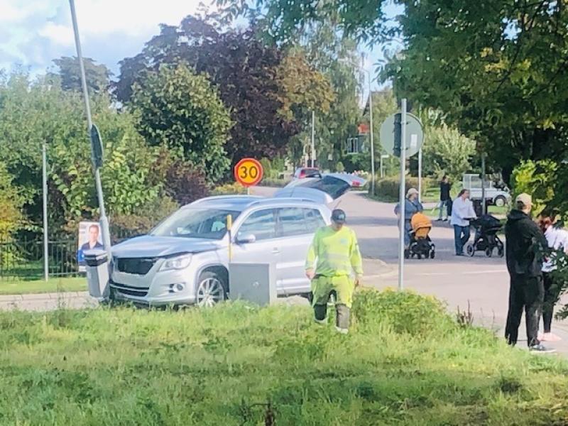 En arbetare i en reflexdräkt står vid en gräsyta nära en silverfärgad SUV som står parkerad vid vägkanten. Människor med barnvagnar och barn går förbi i närheten. En hastighetsbegränsningsskylt på 30 km/h syns.