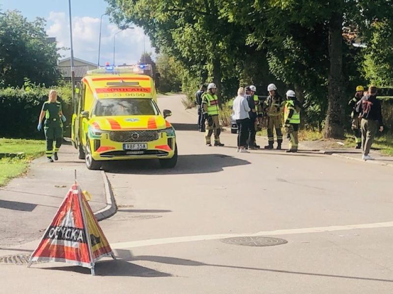 En ambulans och räddningspersonal befinner sig på platsen för en händelse på en bostadsgata, där det står en varningsskylt med texten ”Olycka” framför.