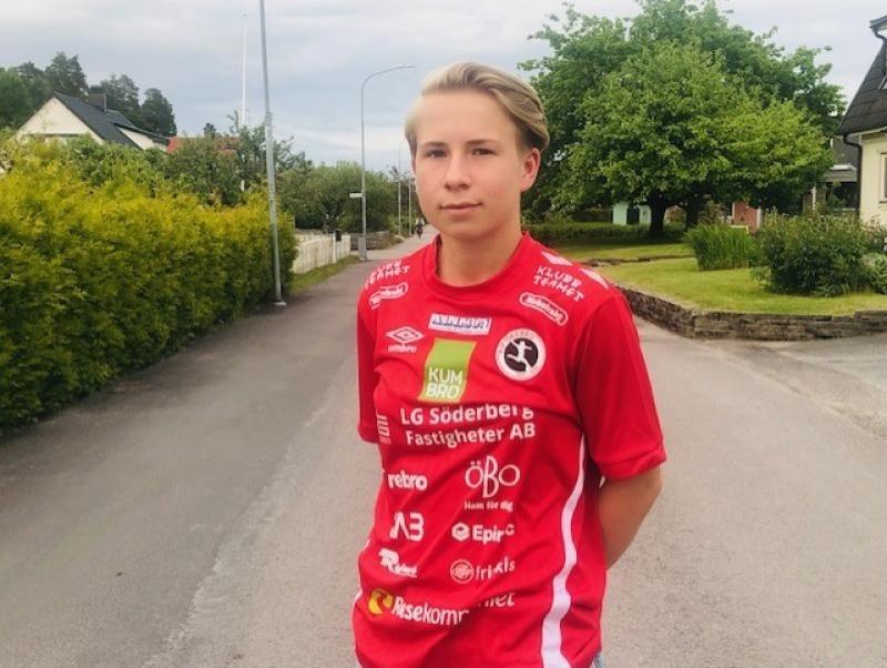 En person står på en bostadsgata iklädd en röd sporttröja med olika sponsorlogotyper. Träd och hus syns i bakgrunden.