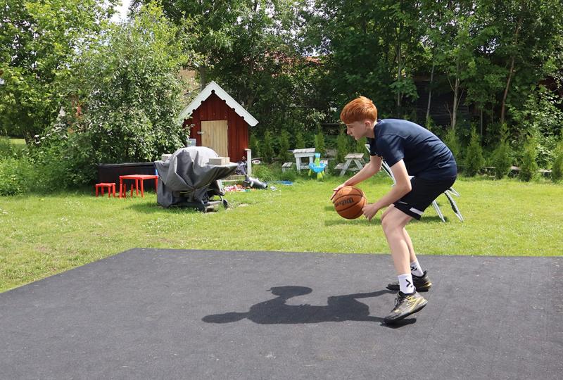 En pojke med rött hår dribblar en basketboll på en utomhusplan i en gräsbevuxen trädgård, med ett litet rött lekhus och utspridda möbler i bakgrunden.