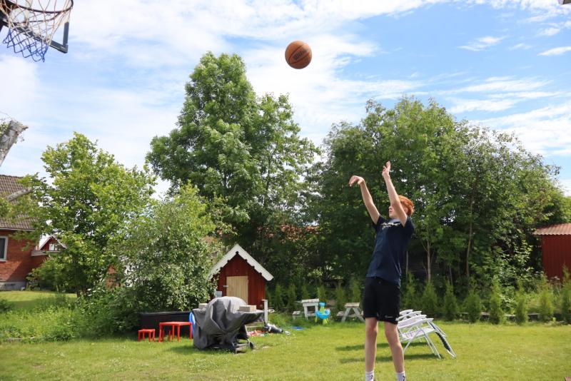En person skjuter en basketboll mot en korg i en gräsbevuxen trädgård med träd och ett litet lekhus i bakgrunden.