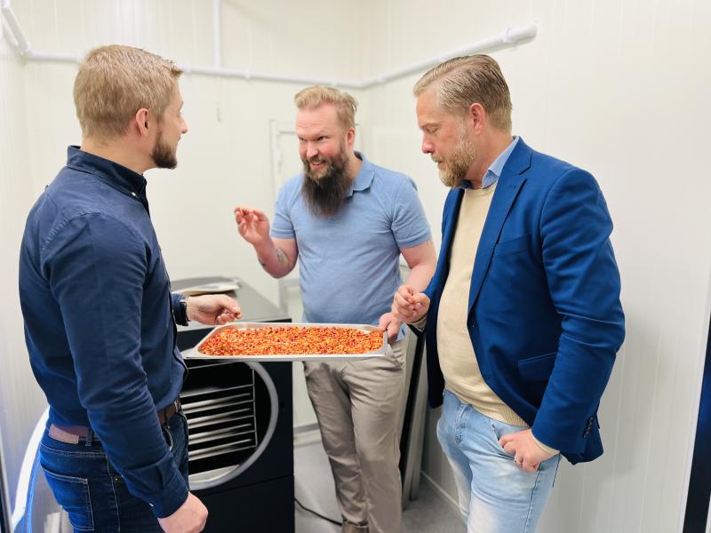 Tre män står i ett rum, en håller en bricka med mat täckt med röda pålägg, medan de andra tittar på den och börjar prata.