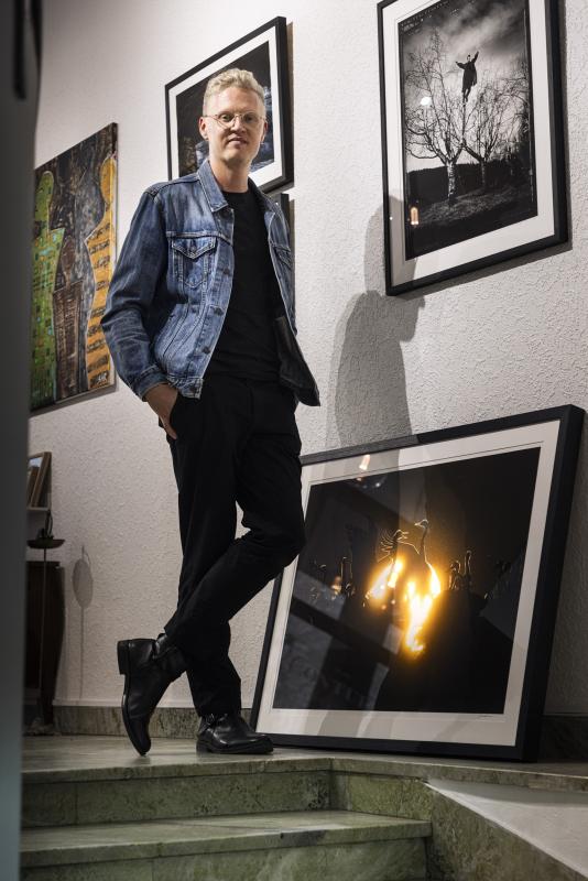 En person i jeansjacka och svarta byxor står på trappor i ett galleri, omgiven av inramade fotografier på vägg och golv.