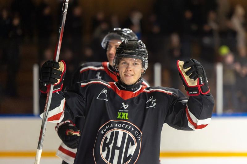 En hockeyspelare i svart uniform ler och gestikulerar med handen på ishallen. En annan spelare syns i bakgrunden.