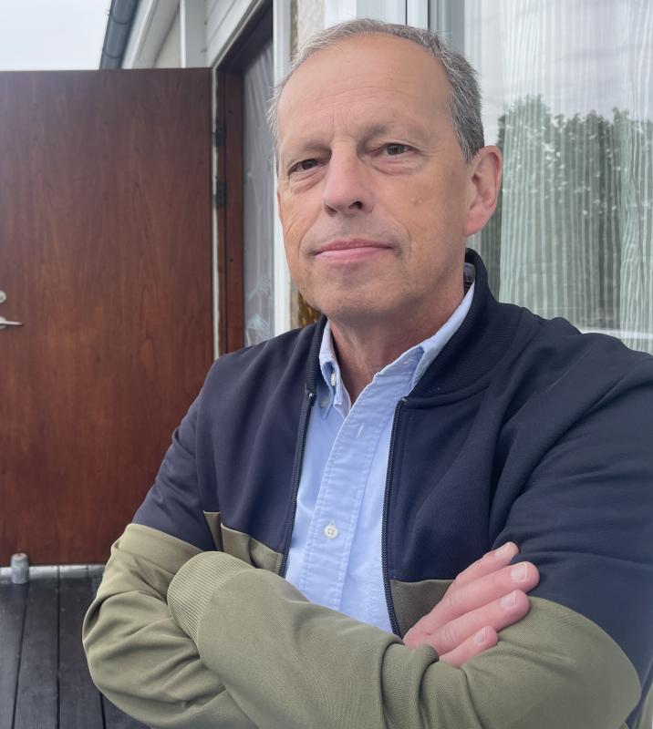 En medelålders man med kort grått hår står utanför med armarna i kors, klädd i ljusblå skjorta och mörk jacka. I bakgrunden syns en trädörr och ett fönster med gardiner.