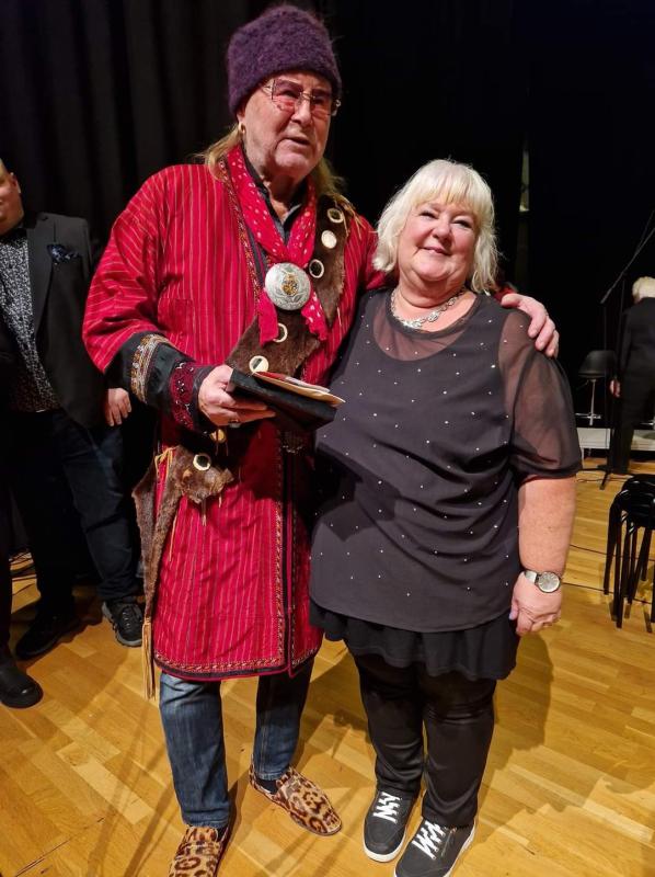 En man i röd mantel och en kvinna i svart dräkt står tillsammans på ett trägolv; mannen håller i ett pris och bär hatt.