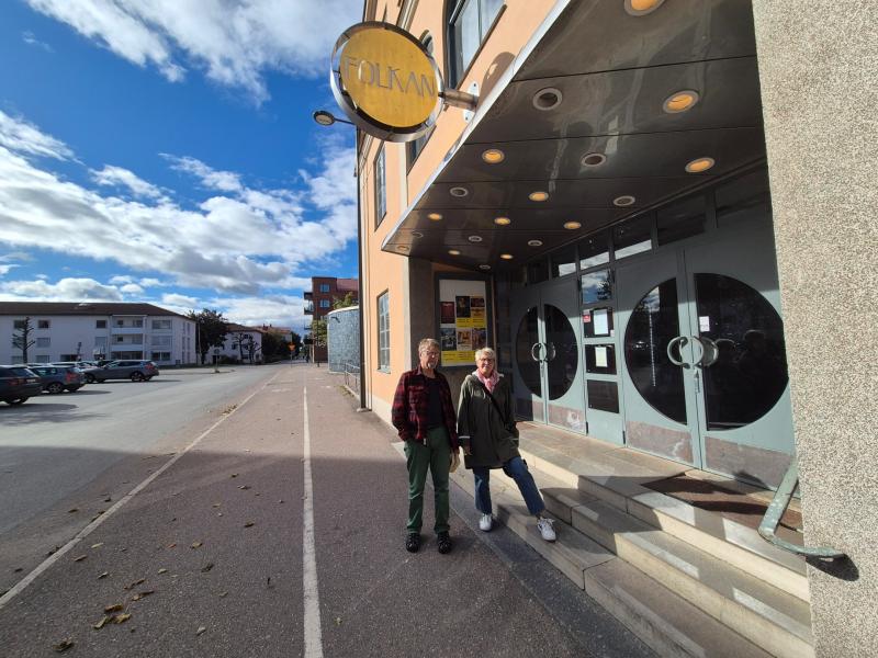 Två personer står utanför en byggnad med en skylt där det står ”Folkana”. Byggnaden har glasdörrar och runda fönster. Gatan bredvid dem är tom under en delvis molnig himmel.