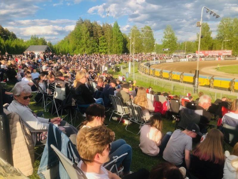 En stor folkmassa sitter i stolar på en gräsbevuxen sluttning och tittar på ett speedwaylopp på en utomhusarena en solig dag.