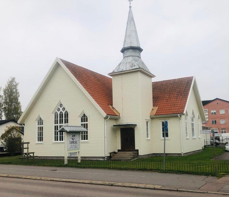 En ljusgul kyrkobyggnad med rött tak, vita lister och ett högt torn står vid en gata, med en skylt med texten 