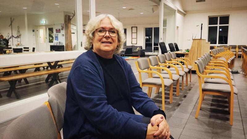 En äldre person med glasögon och grått hår sitter på en stol i ett rum med rader av tomma stolar och bord, vänd mot kameran och ler.