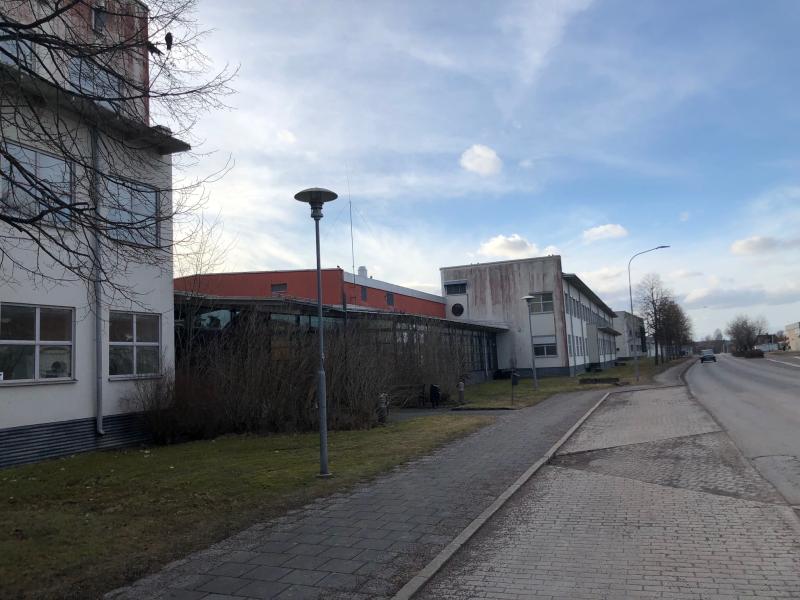 En lugn gata går förbi industribyggnader med stora fönster, en trottoar, gatlyktor och glesa träd under en delvis molnig himmel.