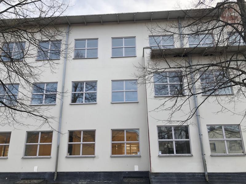 En vit trevåningsbyggnad med flera fönster och lövlösa trädgrenar framför, fotograferad från marknivå.