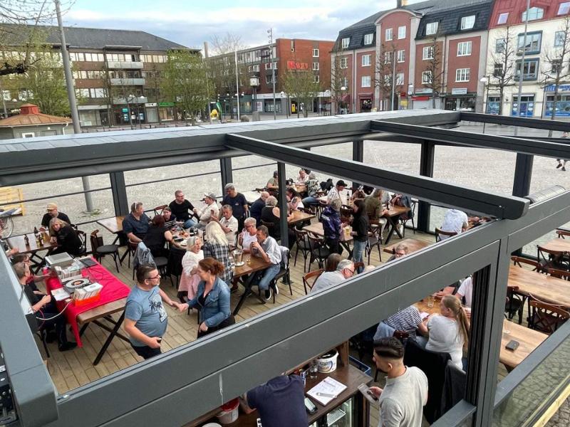 Utomhusmatplats på en terrass med flera våningar med gäster som sitter vid bord och deltar i samtal och middagar, i en stadsmiljö med byggnader i bakgrunden.