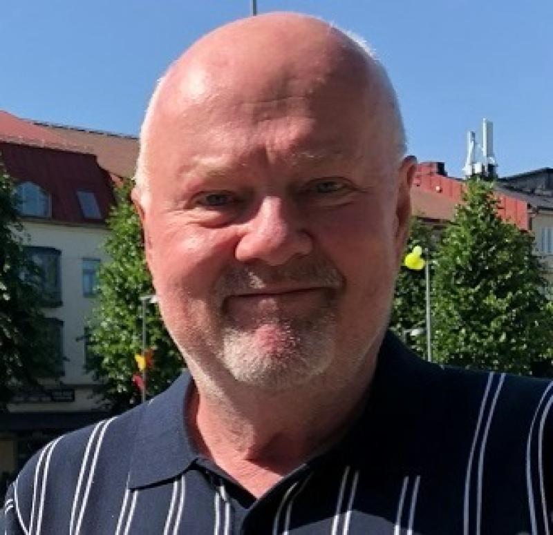 En medelålders man med skalligt huvud och grått skägg, klädd i en mörk randig polotröja, står utomhus en solig dag med byggnader och träd i bakgrunden.