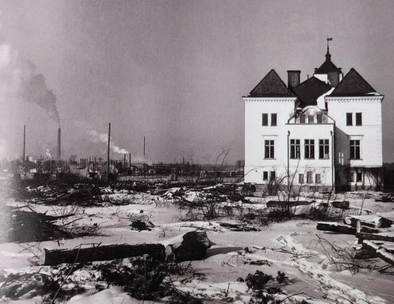 En vit byggnad står ensam i ett snötäckt, öde landskap med skorstenar och industriella ruiner i bakgrunden.