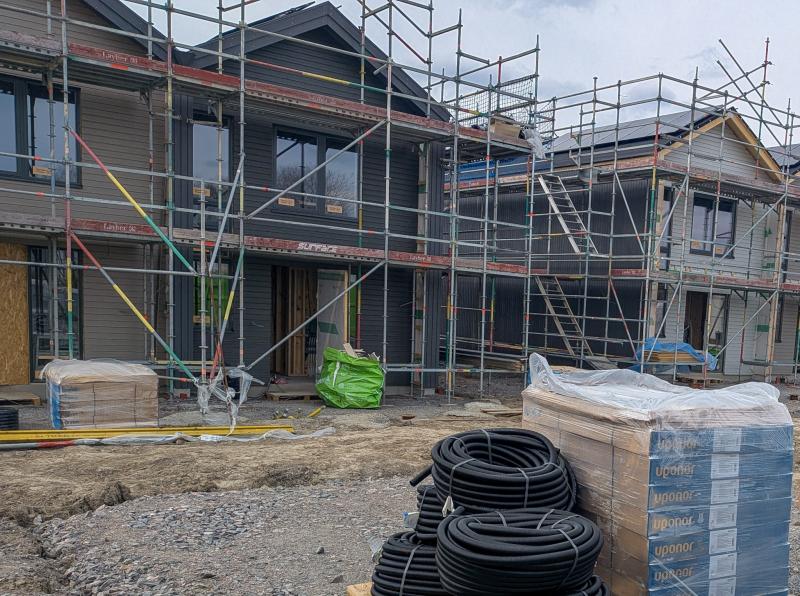 Två delvis uppförda hus med byggnadsställningar, staplar av byggmaterial och rörledningar på en byggarbetsplats.