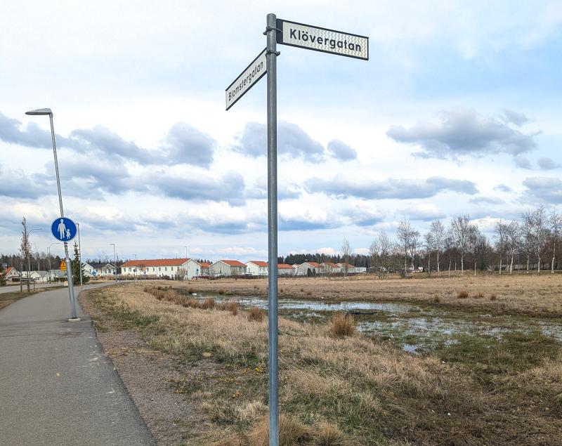 En gatuskylt vid korsningen mellan Klövergatán och en annan väg står bredvid en trottoar och en gräsbevuxen, vattendränkt åker under en molnig himmel.