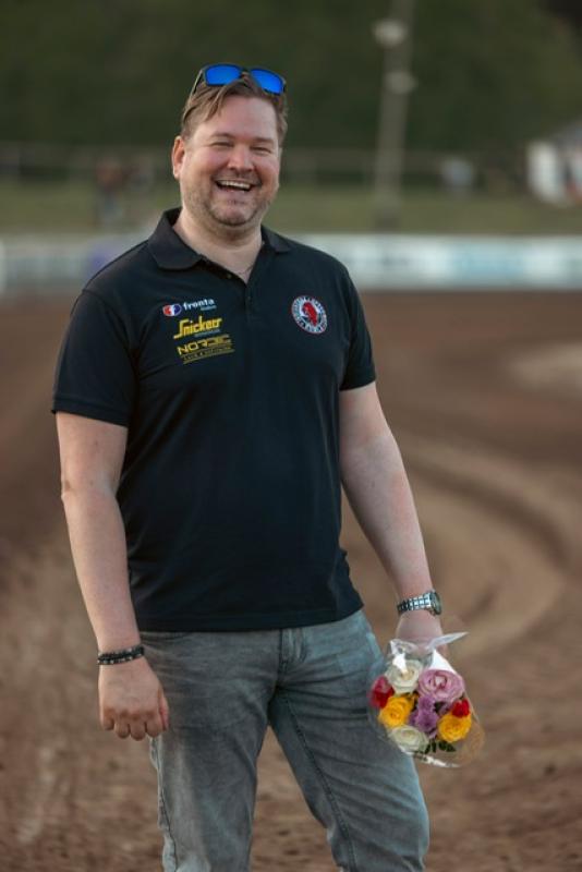 En man i svart pikétröja och jeans står på en grusväg, ler och håller i en bukett färgglada blommor.