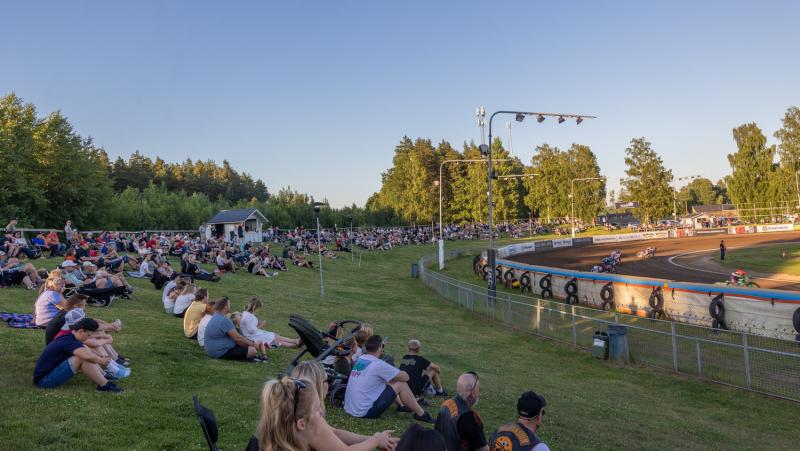 Åskådare sitter på en gräsbevuxen kulle och tittar på en motorcykeltävling på en grusbana under en klar dag, med omgivande träd och en blå himmel i bakgrunden.