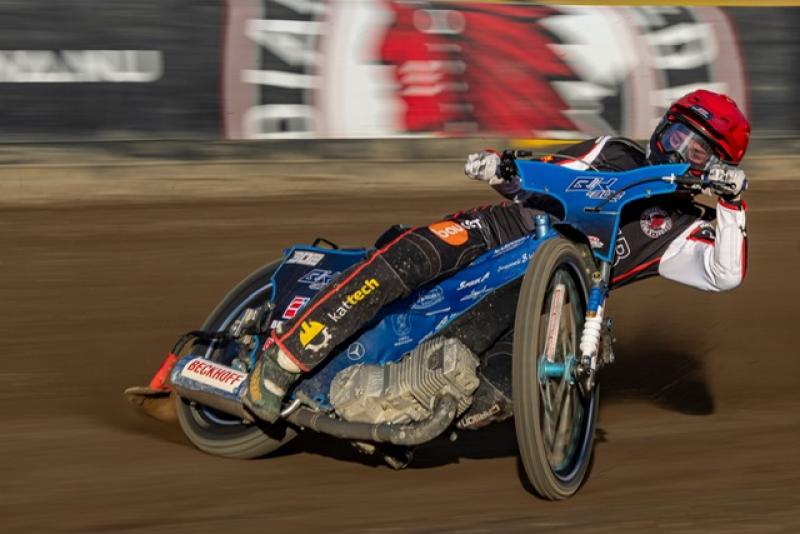 En speedwaymotorcyklist i röd hjälm och racingdräkt lutar sig in i en kurva på en grusbana, med synliga banderoller i den suddiga bakgrunden.