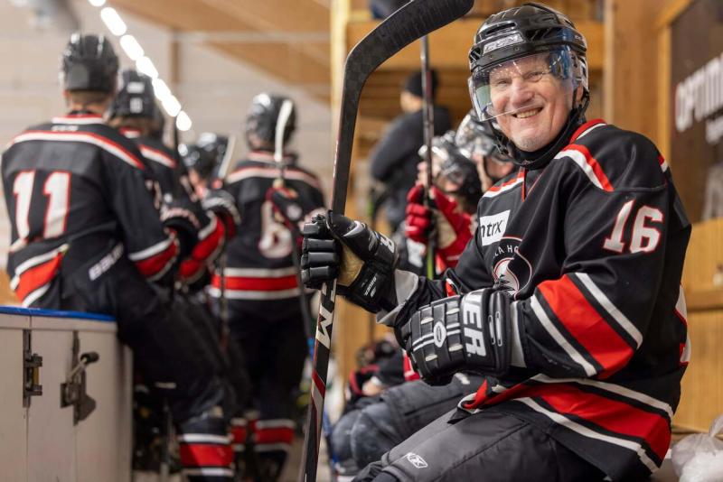 En hockeyspelare i en svart och röd tröja sitter på bänken och ler och håller i en pinne. Andra spelare i liknande uniformer sitter i närheten.