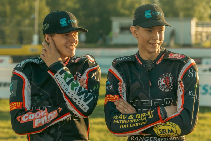 Två motorcyklister i racerutrustning och kepsar ler och står med armarna i kors på en bana. Deras kostymer är märkta med olika sponsorlogotyper.