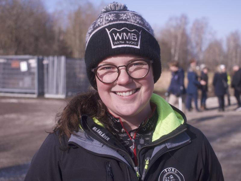 En leende person som bär glasögon, en stickad mössa och en jacka utomhus.