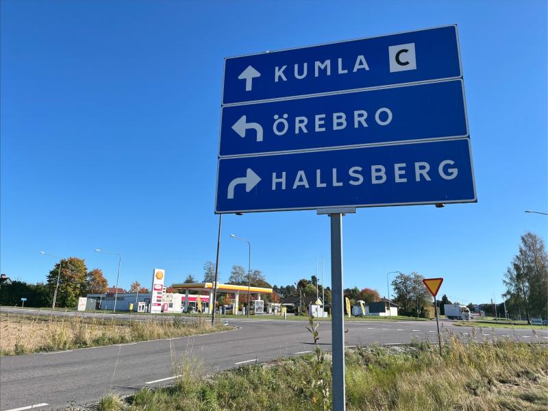 Vägskylt med vägbeskrivning: rakt till Kumla, vänster till Örebro, och höger till Hallsberg, med en bensinmack i bakgrunden.