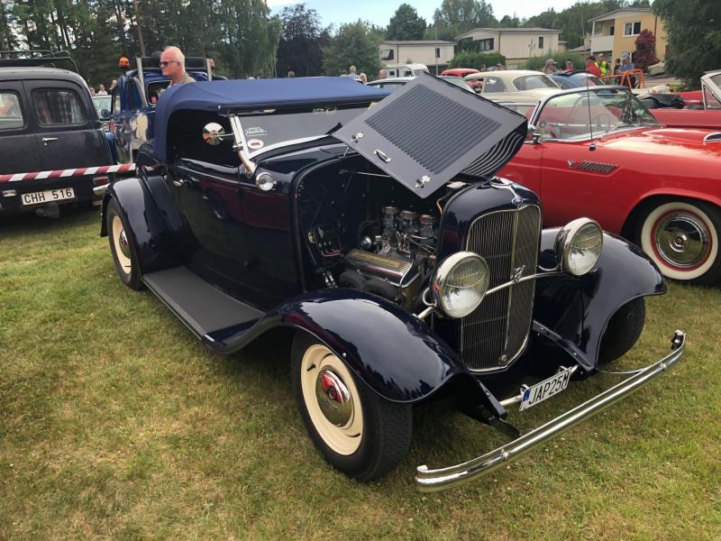 En vintage-cabriolet i marinblått med öppet tak, där motorn syns, står parkerad på gräset vid en veteranbilsutställning med andra bilar och människor i bakgrunden.