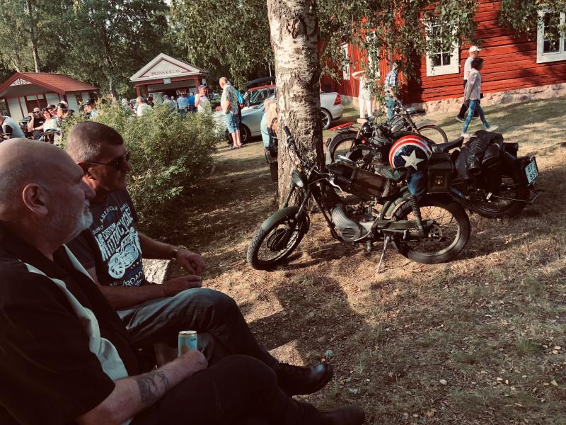 Två män sitter på en bänk bredvid gamla motorcyklar som står parkerade under ett träd vid en utomhussammankomst, med människor och röda träbyggnader i bakgrunden.